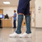 A realistic, square photograph focusing on the lower legs and feet of a nurse standing in a hospital corridor. The nurse wears navy blue scrub pants cuffed above the ankle, knee-high compression socks with a teal background and a white pattern of stethoscopes, hearts, and medical symbols, and clean white nursing clogs. A blurred figure of another healthcare worker is visible at a desk in the background.